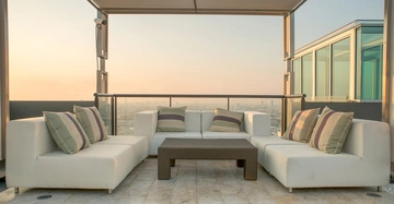 Weiße Sofas mit gestreiften Kissen stehen um einen niedrigen Couchtisch auf einer Terrasse mit Dach in einem Hochhaus mit Stadtblick bei Sonnenuntergang