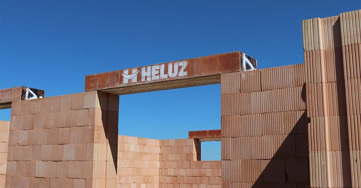 Galerie-Bild 1: Baustelle mit unvollständigen Ziegelwänden unter blauem Himmel ein Stahlbalken mit der Aufschrift HELUZ spannt sich über eine Öffnung in einer Wand