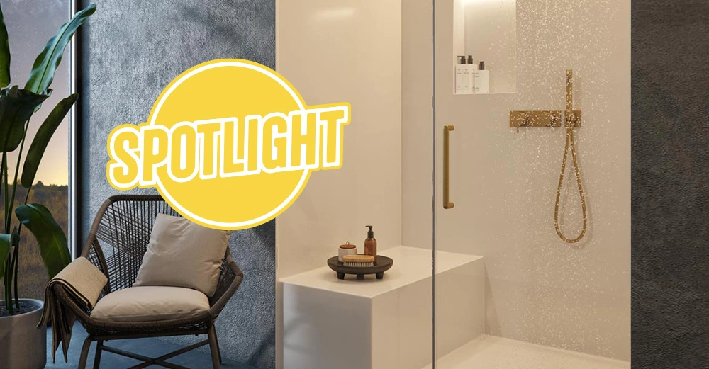 fugenlose, beige Dusche mit goldenen Armaturen, im Vordergrund durchsichtige Glaswand mit Wassertropfen, links ein gemütlicher Sessel mit Sitzkissen.