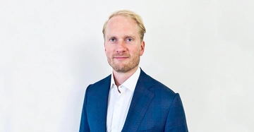 Mann steht vor einem weißen Hintergrund trägt ein blaues Jackett und ein weißes Hemd blickt in die Kamera