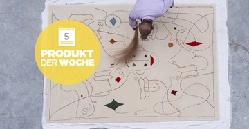 Ein Mann kehrt einen beigen Teppich mit abstrakten Zeichnungen auf grauem Boden. Text im gelben Kreis lautet Produkt der Woche.