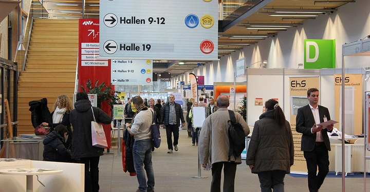 Galerie-Bild 8: Menschen laufen durch eine Messehalle mit Ständen auf beiden Seiten und einer großen Wegweiser-Tafel, die zu den Hallen 1-8, 9-12 und 19 führt.