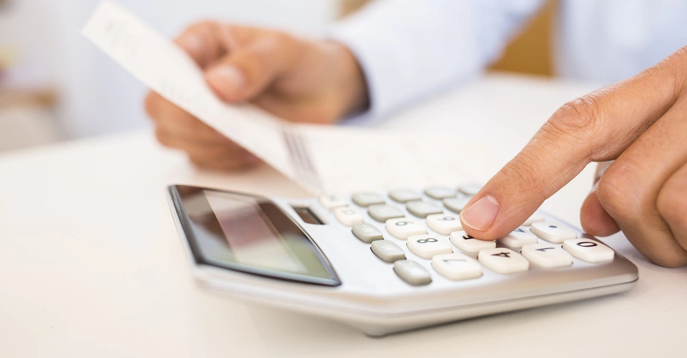 Ein Taschenrechner wird von einer Hand bedient, während die andere Hand einen Beleg hält. Im Hintergrund ist ein unscharfer Raum zu sehen.