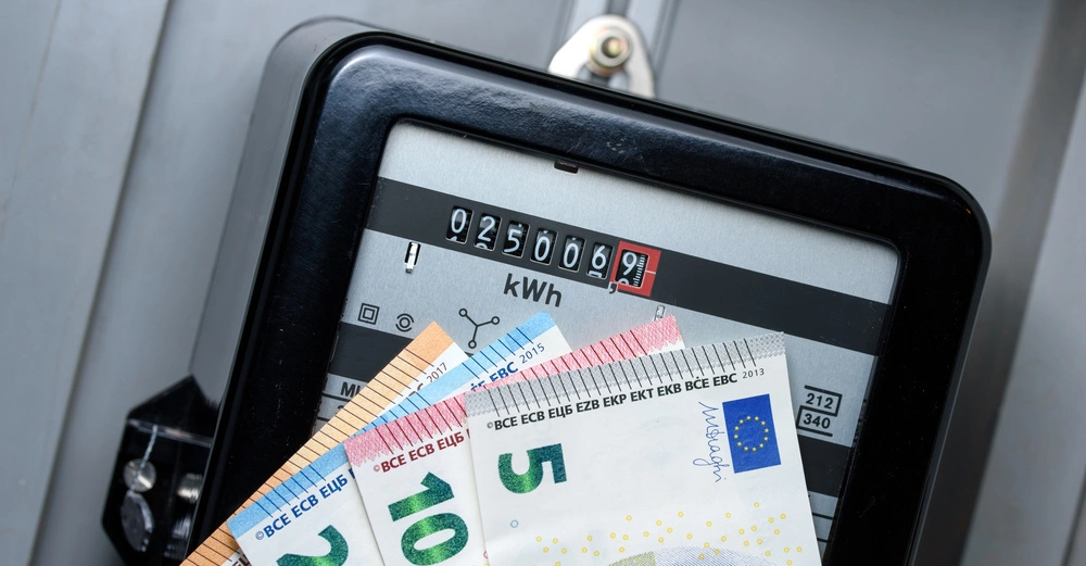 Elektrizitätszähler zeigt 0250069 kWh-Zählerstand an gehaltene Euro-Banknoten davor um Kosten zu symbolisieren, Zähler befindet sich in einem Metallgehäuse.