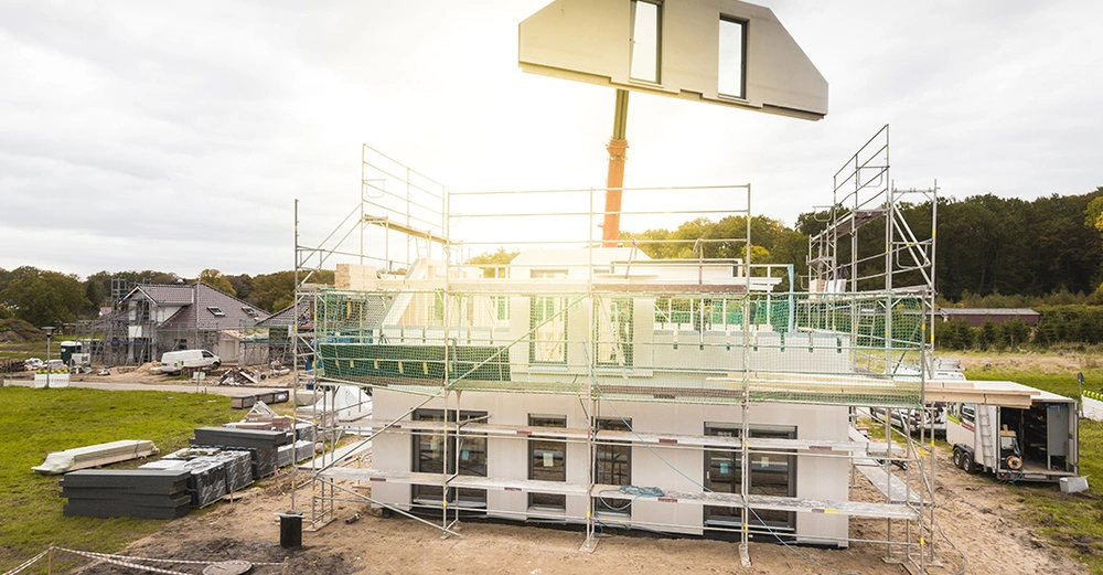 Kran liefert Hausteil mit Fensteröffnung von einem Fertighaus auf die Baustelle, wo schon ein Teil des Erdgeschoßes des Fertighauses steht.