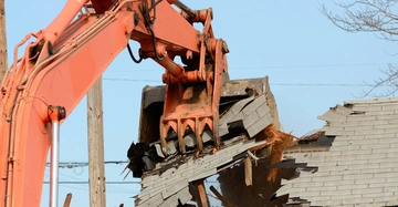 Ein großer, orangener Bagger demoliert das Dach eines Gebäudes im Freien, umgeben von Strommasten und vereinzelten Bäumen bei klarem Himmel.