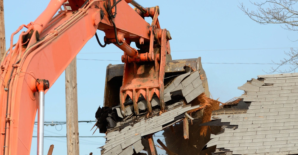 Ein großer, orangener Bagger demoliert das Dach eines Gebäudes im Freien, umgeben von Strommasten und vereinzelten Bäumen bei klarem Himmel.