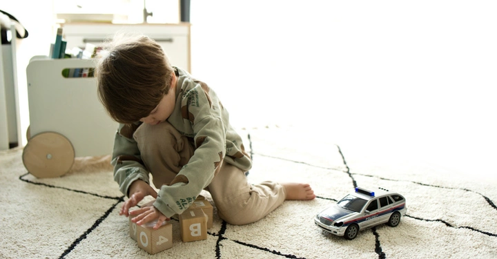 Ein Kind sitzt im Schneidersitz auf einem Teppich und spielt mit Holzwürfeln. Ein Spielzeug-Polizeiauto steht daneben. Im Hintergrund steht ein Spielzeugwagen.