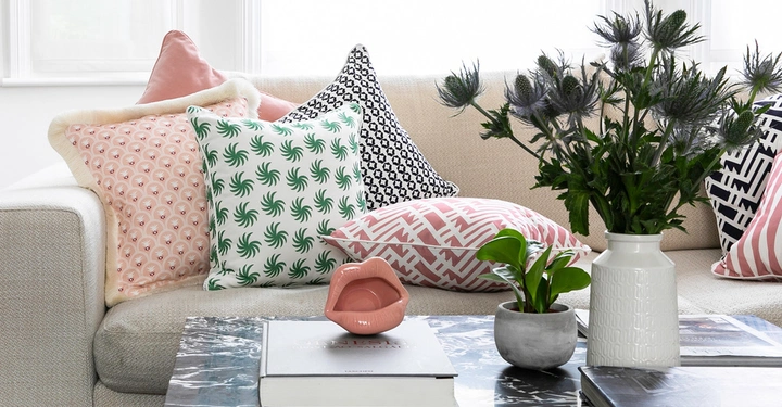 Mehrere gemusterte Kissen liegen dekorativ auf einem beigen Sofa in einem hellen Wohnzimmer. Auf einem Marmortisch davor stehen stilisierte Lippenfigur Pflanzen und Bücher.