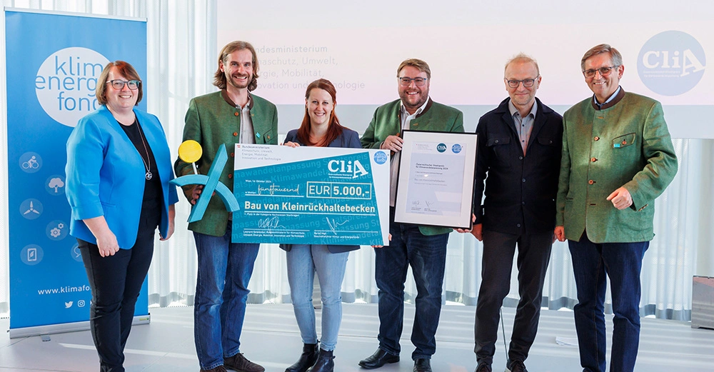 Die 6 Preisträger des CliA Staatspreises für Nachhaltigkeit stehen auf blau-weißer Bühne und lächeln mit ihren Auszeichnungen in die Kamera. 