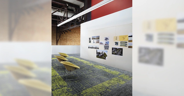 Galerie-Bild 8: Vier gelbe Stühle stehen in einem Raum mit grauem und grünem Teppichboden vor einer Wand mit ausgestellten Fotos und Zeichnungen in einem industriellen, offenen Büro.