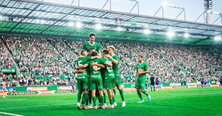 Galerie-Bild 1: Fußballspieler umarmen sich auf einem Fußballfeld, umgeben von jubelnden Zuschauern in einem vollbesetzten Stadion. Die Spieler tragen grüne Trikots mit Logos wie Allianz RHIEM ENERGIE auf der Vorderseite.