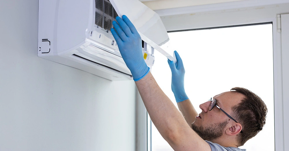 Techniker wartet wandmontierte Klimaanlage in hellem Raum mit großen Fenstern. Reinigung des Filters mit blauen Handschuhen