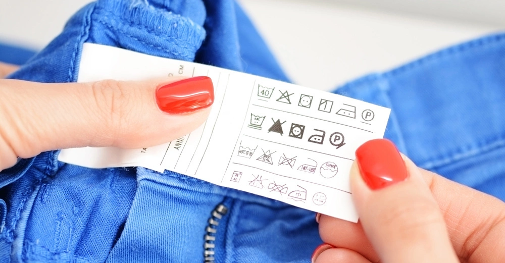 Eine Hand mit roten Nägeln hält ein Etikett mit Pflegehinweisen aus einem blauen Kleidungsstück Hintergrund unscharf Etikett zeigt verschiedene Pflege- und Waschsymbole