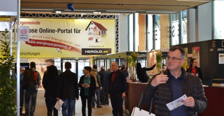 Galerie-Bild 31: Mann hält Papiere in der Hand gehend. Menschen gehen und stehen im Messezentrum. Banner mit Text: Kostenlose Online-Portal für Ihre Energiespargemeinde www.energiespargemeinde.at. Umgebung modern mit Informationsschalter.