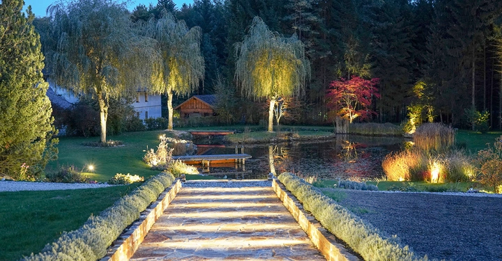 Beleuchteter Steinweg führt zu einem Teich mit Holzstegen in einem weitläufigen Garten mit Bäumen und Büschen bei Dämmerung umgeben von Wald und Gebäuden.