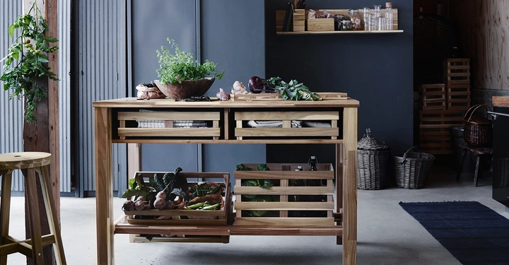 Holztisch hält frische Gemüse und Küchenutensilien. Schüsseln mit Pflanzen und Gemüse liegen auf dem Tisch. In einem gut organisierten Küchenraum mit grauen Schränken und Regalen.