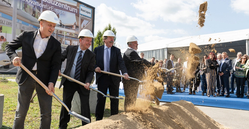 Mehrere Männer mit Helmen heben mit Schaufeln Sand während einer feierlichen Grundsteinlegung in die Luft im Freien vor einem großen Plakat mit der Aufschrift Neues Blaue Lagune Bauzentrum Das Innovationszentrum für Europa