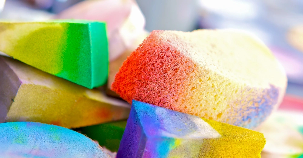 Bunte Schwämme liegen übereinander nah beieinander auf einem unscharfen Hintergrund.