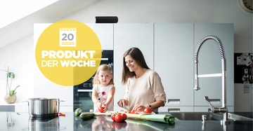 Eine Frau schneidet Gemüse auf der Küchenarbeitsplatte während ein kleines Mädchen zuschaut. Im Hintergrund helle Schränke. Text: 20 PRODUKT DER WOCHE