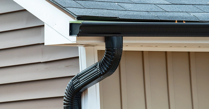 Schwarze Kastenrinne mit passendem Fallrohr, installiert an einem Haus mit Schindeldach und heller Fassade.