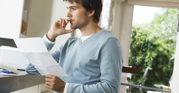 Mann sitzt und liest ein Papier in einem modernen, gut beleuchteten Raum mit großen Fenstern und grüner Vegetation im Hintergrund, während er nachdenklich auf seine Finger beißt.