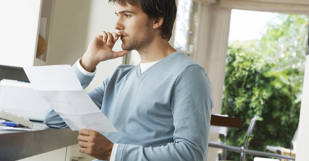 Mann sitzt und liest ein Papier in einem modernen, gut beleuchteten Raum mit großen Fenstern und grüner Vegetation im Hintergrund, während er nachdenklich auf seine Finger beißt.