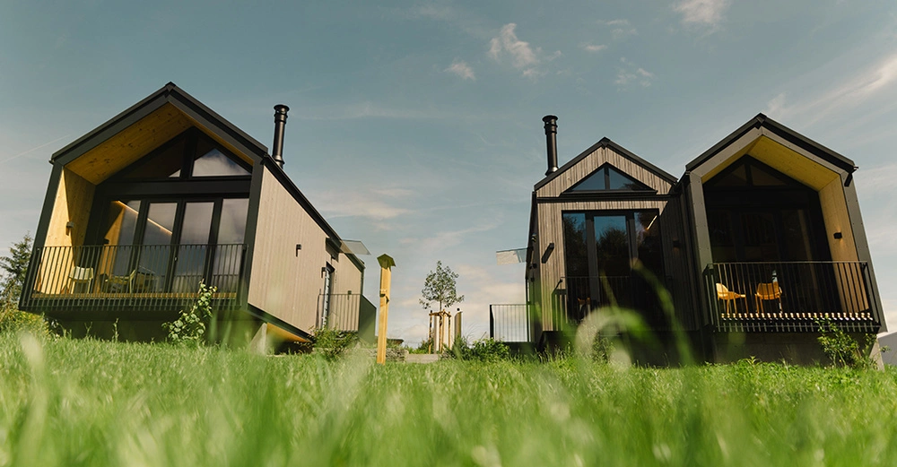 Aufnahme von drei Tiny Houses auf grüner Wiese vor blauem Himmel. Im Vordergrund saftiges grünes Gras. Häuser aus dunklem Holz mit großen Fensterfronten und Loggien. 