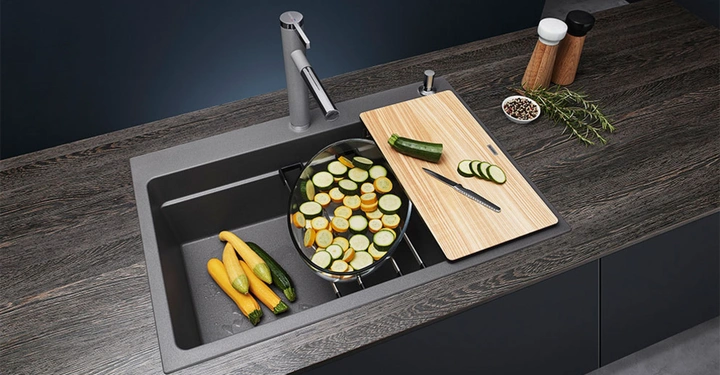 Gemüse wird auf einem Schneidebrett neben einer grauen Spüle geschnitten. In der Spüle befinden sich geschnittene Zucchinischeiben in einem Sieb. Der Hintergrund ist eine dunkle Arbeitsplatte mit Gewürzen und Kräutern.