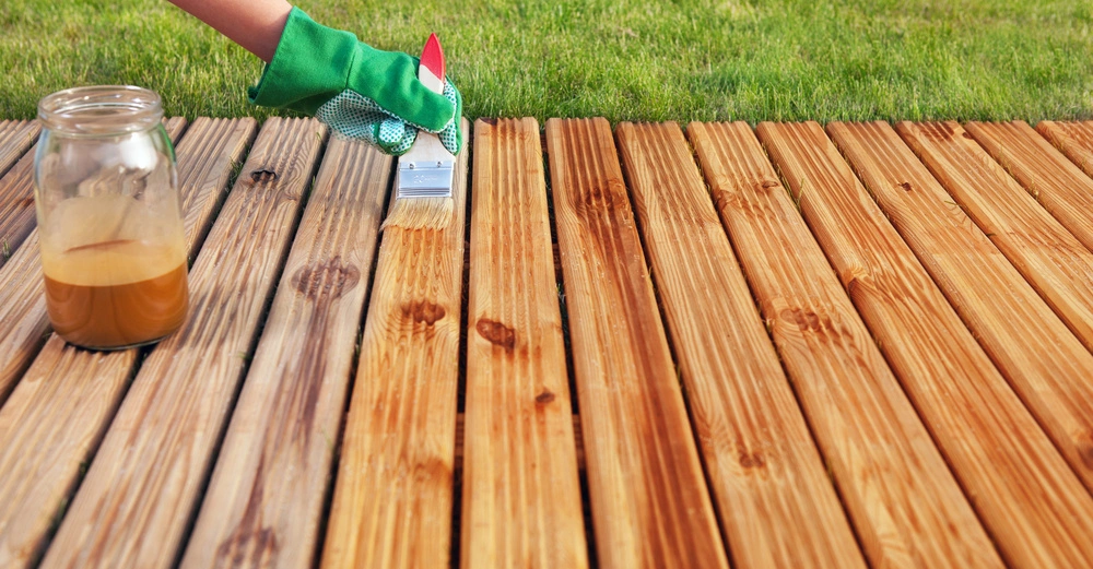 Hand in grünem Handschuh malt Holzbretter auf Terrasse mit Pinsel und Glas Behälter auf Wiese im Freien.