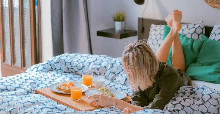 Person liegt auf dem Bett und frühstückt Tablett mit Frühstück Essen auf dem Bett Leichter Raum mit Pflanzen und Türen im Hintergrund Kissen mit geometrischen Mustern und grünem Kissen.