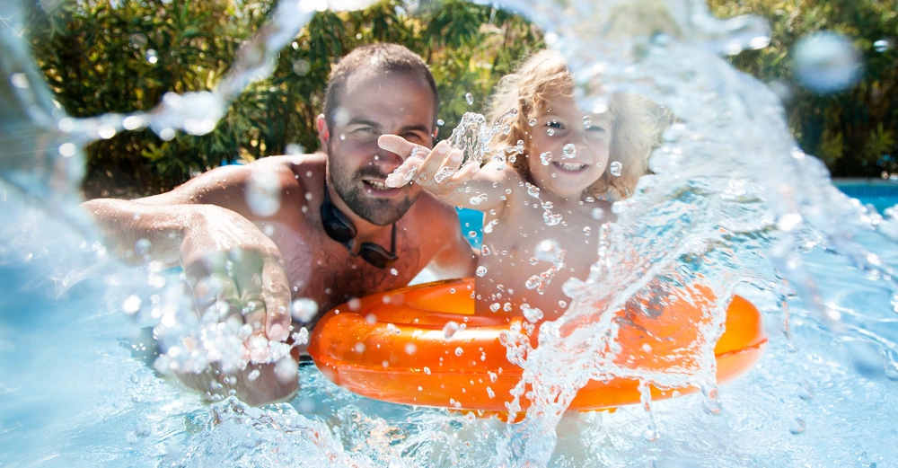 Erwachsener und Kind spielen mit einem orangefarbenen Schwimmring im Wasser Outdoor Pool mit spritzendem Wasser und grünem Laub im Hintergrund bei sonnigem Wetter