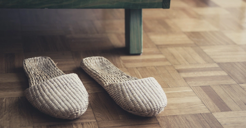 Geflochtene Hausschuhe liegen auf einem Parkettboden neben einem grünen Möbelstück in einem Innenraum.