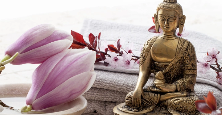 Goldene Buddha-Statue sitzt meditierend auf einem Tisch umgeben von rosa Blütenzweigen und weißen Handtüchern im Hintergrund helle Umgebung
