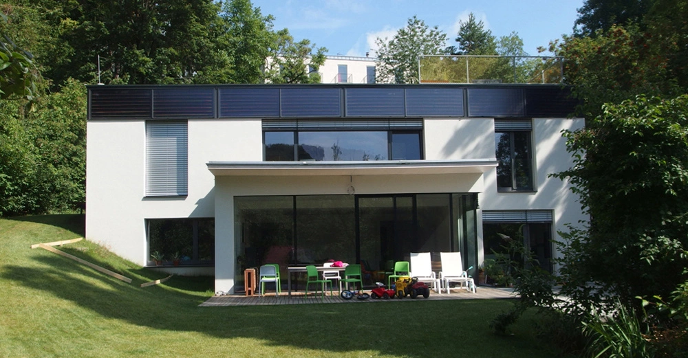 Ein modernes zweistöckiges Haus steht in einem gepflegten, grünen Garten. Auf der Terrasse vor dem Haus befinden sich bunte Stühle, Spielzeuge und Möbel. Bäume umgeben das Haus im Hintergrund.