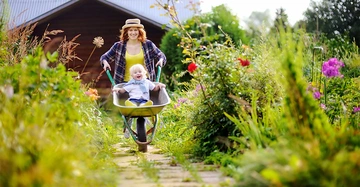 Eine Frau schiebt eine Schubkarre mit einem lachenden Kind durch einen blühenden Gartenweg. Im Hintergrund ein Holzhäuschen.