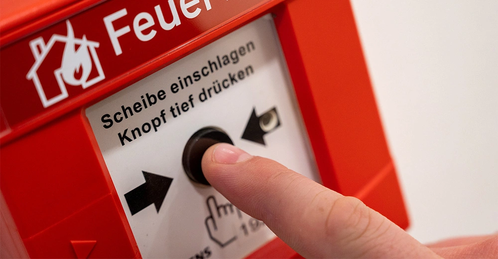 Nahaufnahme eines Feuermelders mit Finger, der den schwarzen Auslöser betätigt.