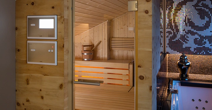 Sauna-Holzkammer mit geöffneter Glastür zeigt Inneneinrichtung mit Holzbänken und einem Holzeimer außerhalb der Sauna ist ein sprudelnder Whirlpool zu sehen mit Mosaikfliesen an der Wand