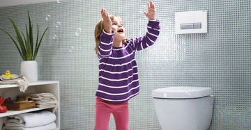 Ein Kind in einem gestreiften Pullover fängt Seifenblasen in einem hellen Badezimmer mit grün-weißer Fliesenwand auf einem flauschigen Teppich neben einem wandmontierten WC und Regalen mit Handtüchern und Pflanzen.