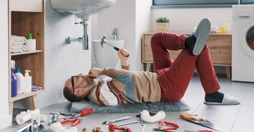 Mann in Hauspatschen liegt auf Rücken unter Waschbecken im Badezimmer und versucht den Abfluss zu reparieren. Sieht hilflos und verwirrt aus. Aber lustig.