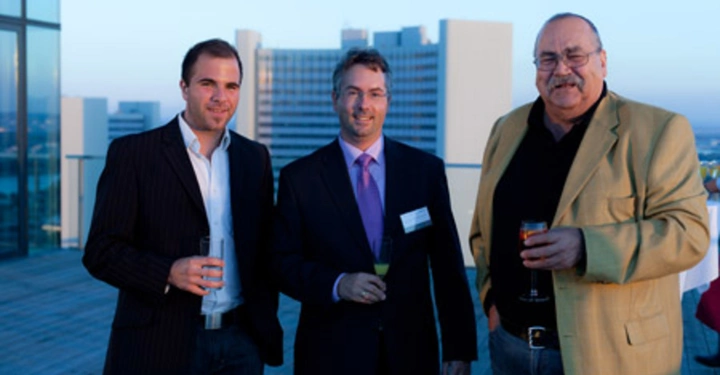 Galerie-Bild 15: Drei Männer halten Getränke in den Händen stehend auf einer Dachterrasse eines hochmodernen Gebäudes bei Sonnenuntergang in einer urbanen Umgebung.