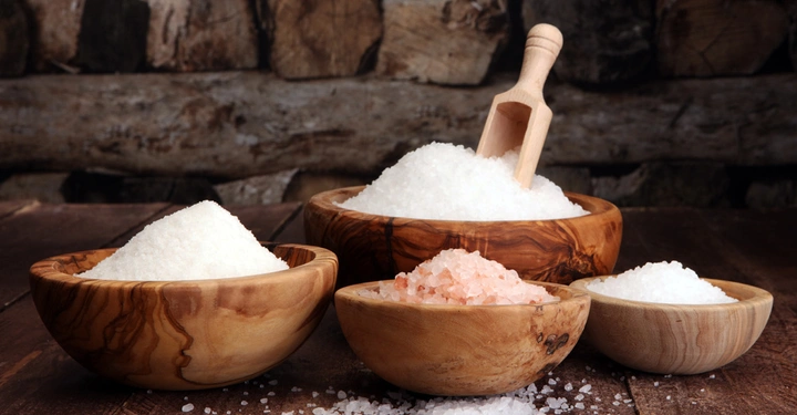 Galerie-Bild 5: Verschiedene Salzsorten liegen in Holzschalen auf einem Holztisch in einer rustikalen Umgebung. Eine hölzerne Schaufel steckt in der größten Schale mit grobem Salz.