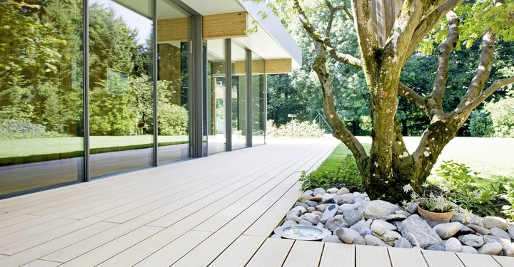 Baum wächst aus Steingarten heraus ruhend neben modernen Glaswänden des Gebäudes Terrasse aus Holzbrettern in einem gepflegten grünen Garten mit Rasen und dichten Bäumen im Hintergrund