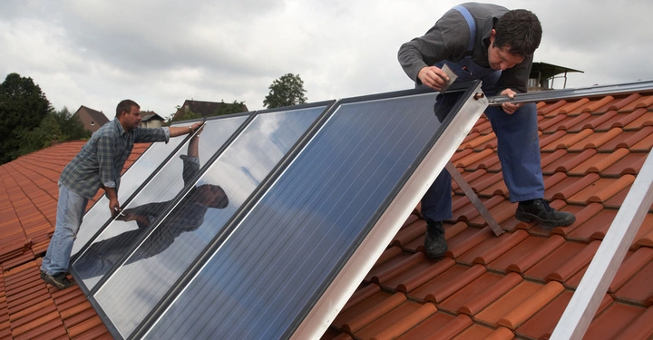 Männer installieren Solarpaneele auf einem Ziegeldach in einer Wohngegend an einem bewölkten Tag. Einer kniet und justiert die Paneele, während der andere sie hält und positioniert.