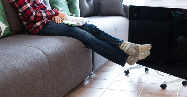 Eine Person liest ein Buch auf einem grauen Sofa. Ihre Beine ruhen auf einem Tisch in einem gut beleuchteten Wohnzimmer mit Pflanzenkissen und Fliesenboden.