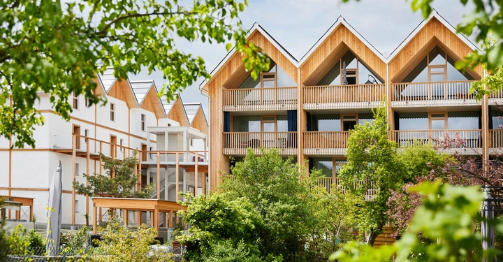 Mehrstöckiges Holzhaus mit Balkonen in anderer Kulisse von kleinen Bäumen und Pflanzen. Neben einem ähnlichen Haus und blauem Himmel. Fenster und Haupttüren sichtbar.