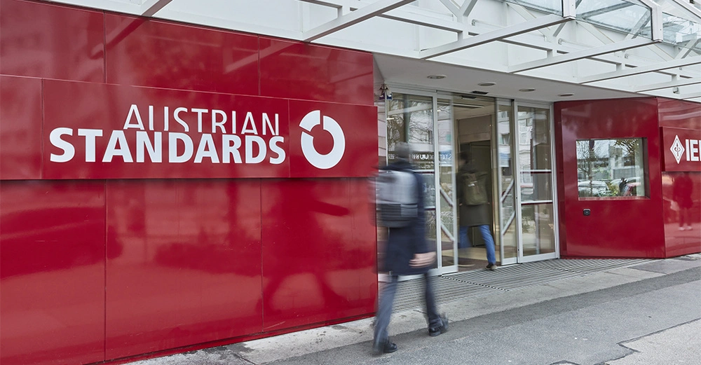 Haupteingang des Austrian Standards Bürogebäudes in Wien, typisch rote Glanzfassade, großer Schriftzug mit Austrian Standards Logo. Eine Person geht vorbei, eine in das Gebäude rein