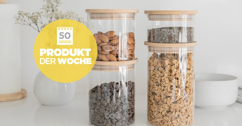 Drei Glasbehälter mit Holzdeckeln beinhalten Mandeln Schokotropfen Müsli und Samen stehen nebeneinander auf einer weißen Oberfläche Kalendericon gelber Kreis Text 50 Produkt der Woche links daneben.