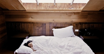 Person schläft zugedeckt in einem weißen Bett in einem holzvertäfelten Raum unter einem Dachfenster.