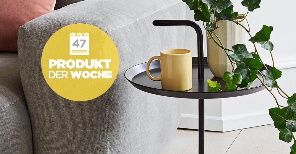 Ein kleiner schwarzer Beistelltisch trägt eine gelbe Tasse und eine Vase mit grünem Laub. Der Tisch steht neben einem grauen Sofa im Wohnzimmer. Text: 47 PRODUKT DER WOCHE